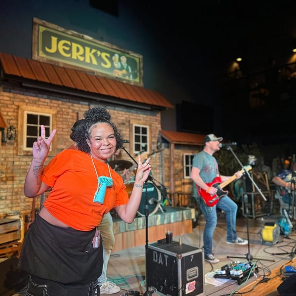 A woman in an orange shirt and black apron smiles, making peace signs in front of a band playing.