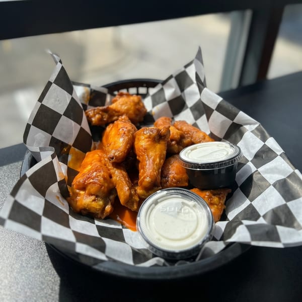 A basket of buffalo chicken wings with two containers of dipping sauce on a checkered paper liner.