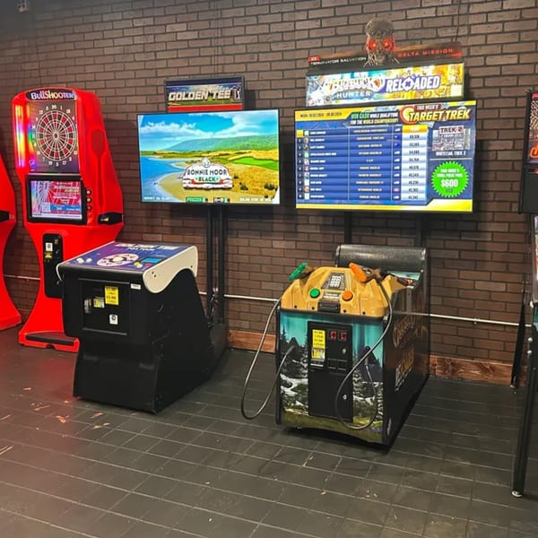 Arcade games, including Bullshooter darts, Golden Tee golf, and Big Buck Hunter, line a brick wall.