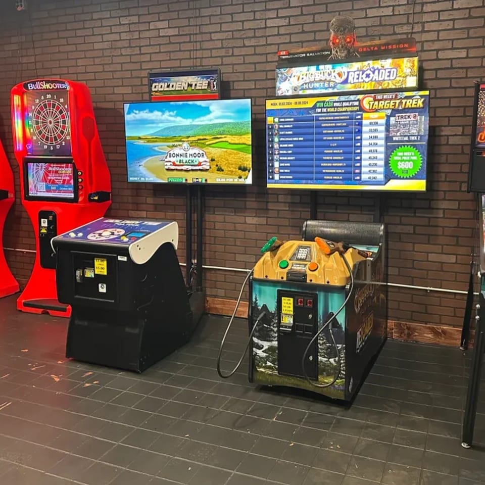 Arcade games, including Bullshooter darts, Golden Tee golf, and Big Buck Hunter, line a brick wall.