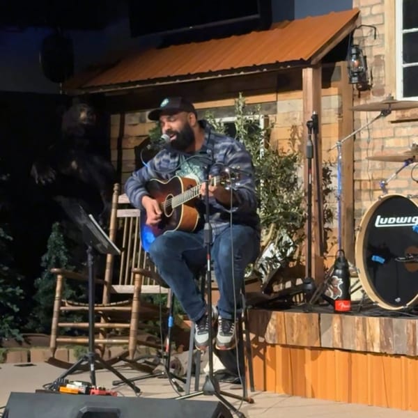 A man with a beard and cap plays an acoustic guitar on a stage with a drum set.