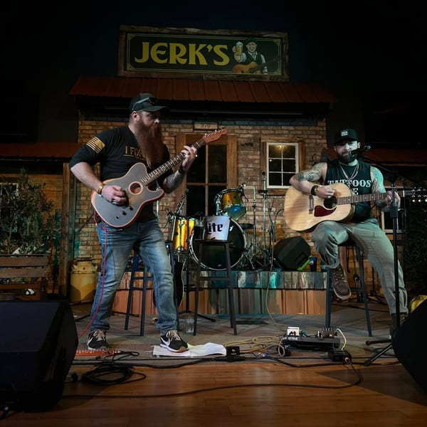 Two musicians playing guitars on stage at Jerk's, with a drum set and a Miller Lite bucket.