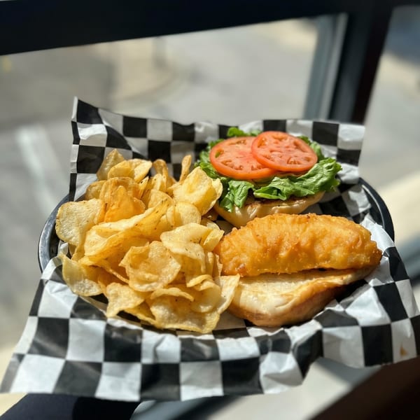 A fried fish sandwich with lettuce and tomato, served with potato chips in a checkered paper-lined basket.