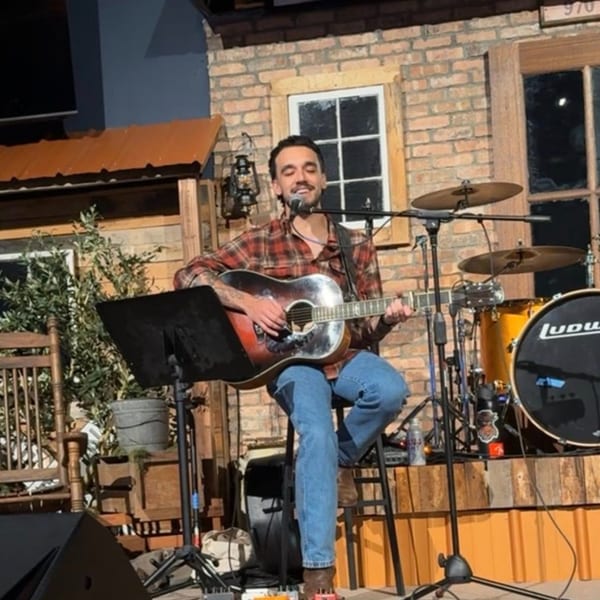 A musician in a plaid shirt plays an acoustic guitar and sings into a microphone on stage.
