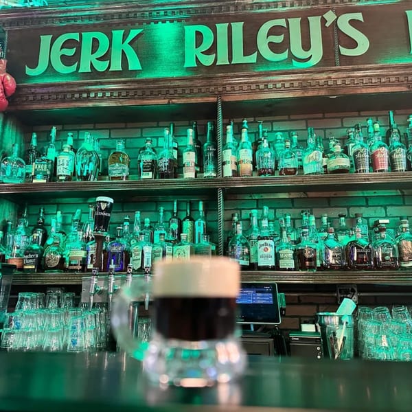 A pint of Guinness sits on a bar counter with shelves of liquor bottles and a "Jerk Riley's" sign.