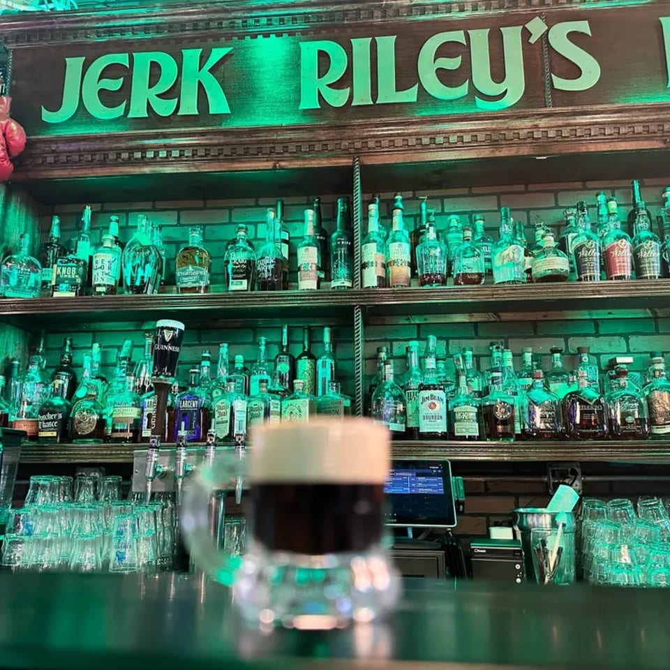 A pint of Guinness sits on a bar counter with shelves of liquor bottles and a "Jerk Riley's" sign.