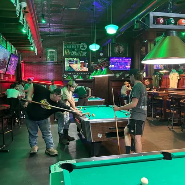 Patrons playing pool in a dimly lit bar with green and red lighting and multiple TVs.