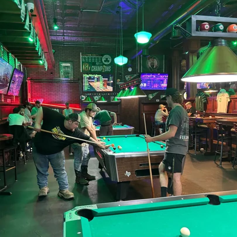 Patrons playing pool in a dimly lit bar with green and red lighting and multiple TVs.