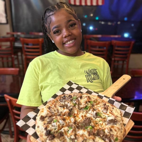 A smiling woman in a neon green shirt holds a large pizza with meat and vegetables.