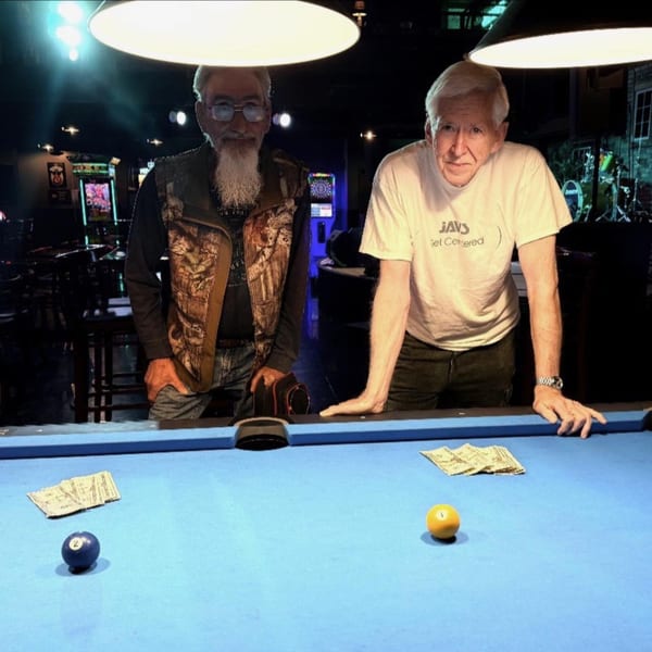 Two men stand over a blue pool table with money and two billiard balls on it.