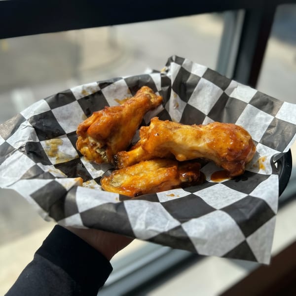 Hand holding a basket of four glazed chicken wings in black and white checkered paper.