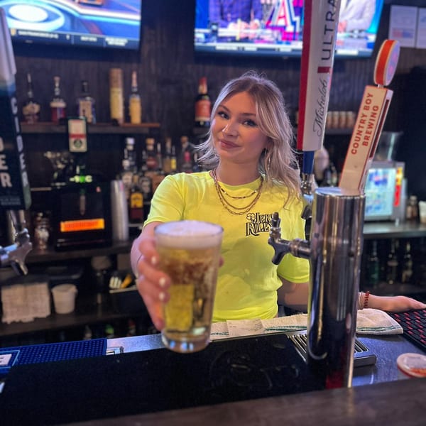 A smiling bartender in a neon yellow shirt serves a glass of beer over a dark bar.
