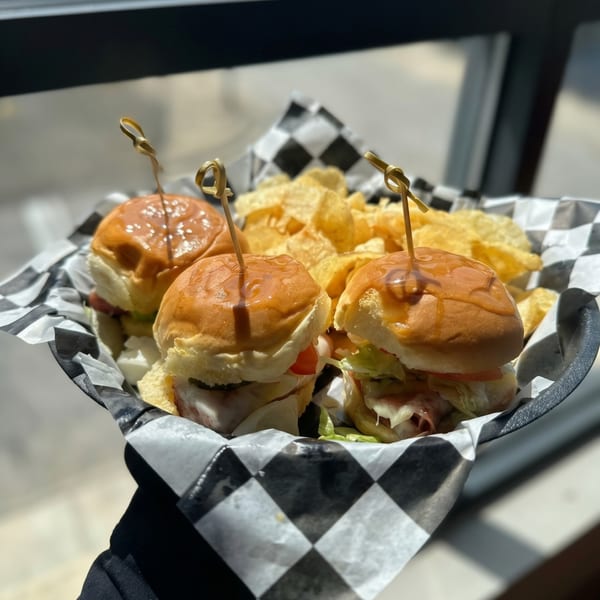 Three sliders with sauce, lettuce, and tomato, served with potato chips in a checkered basket.