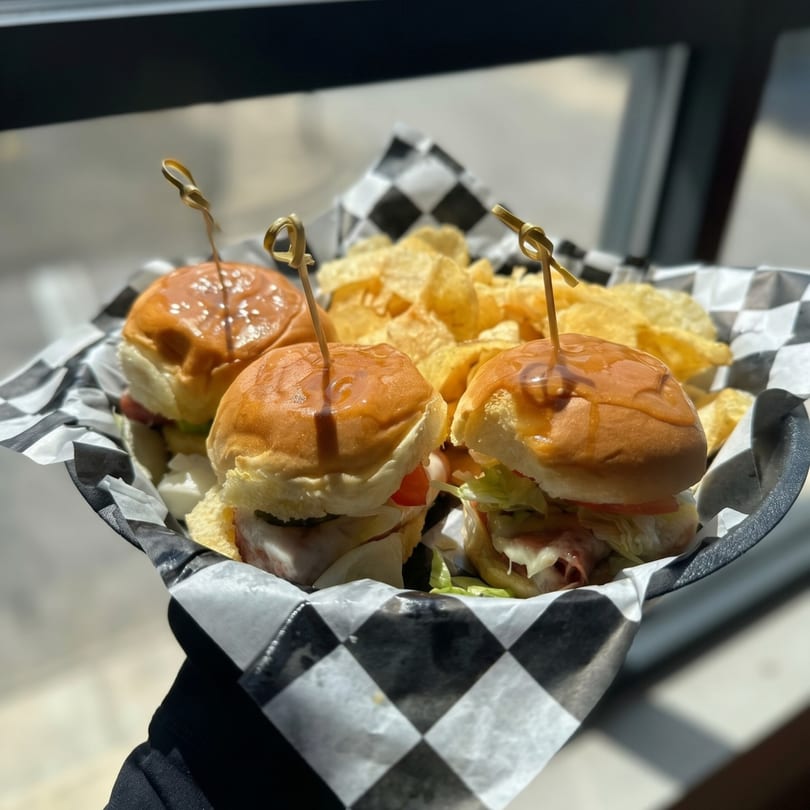 Three sliders with sauce, lettuce, and tomato, served with potato chips in a checkered basket.